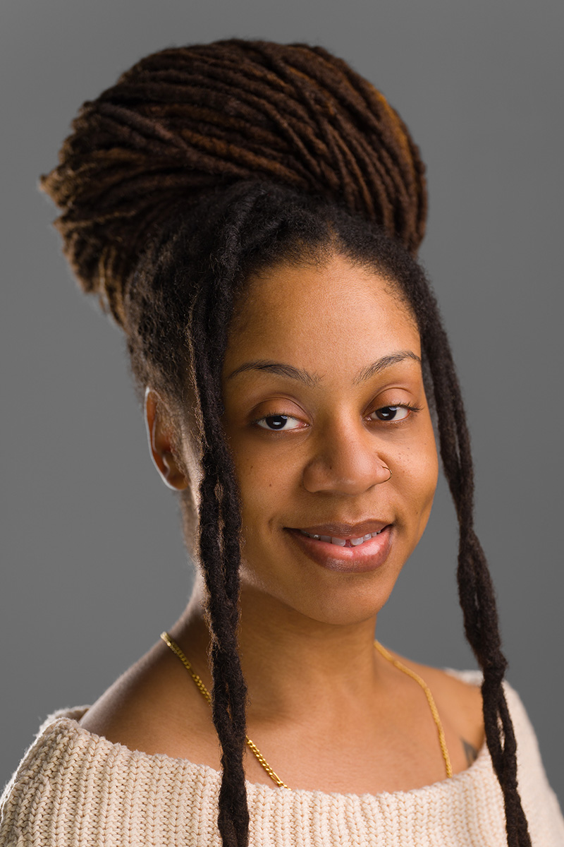 Studio headshot of a woman with locs styled in a high bun against a gray background