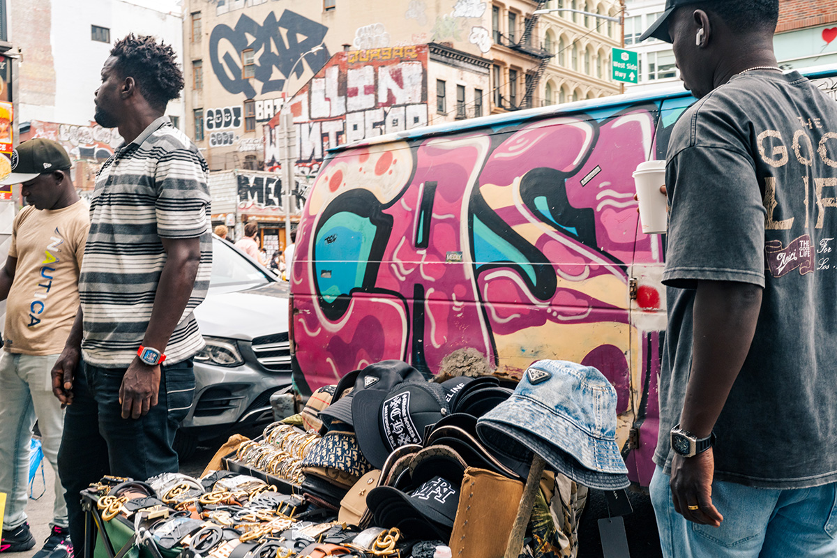 Three street vendors beside a table of hats and belts in front of a pink graffiti-covered van