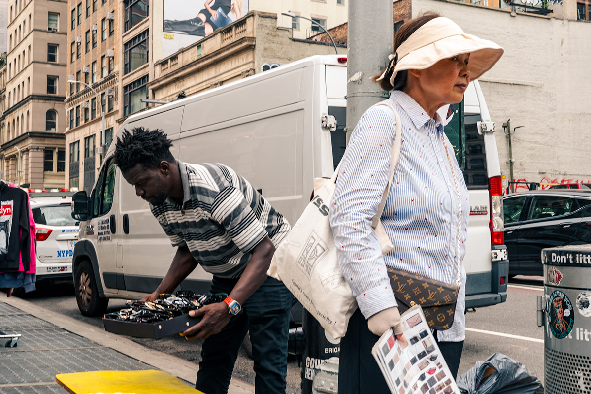 Street vendor arranging merchandise beside a white van as a woman in a wide-brimmed hat walks past