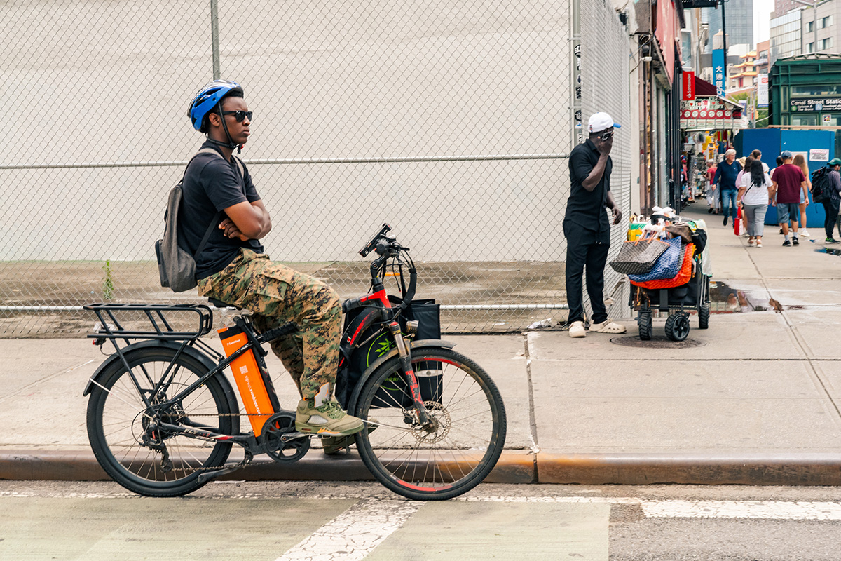 Delivery worker in a blue helmet waiting on an e-bike beside another vendor's cart on Canal Street
