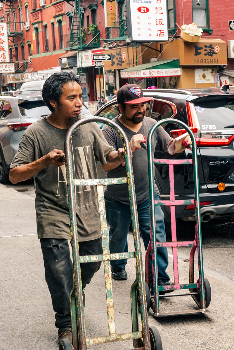 Two workers pushing empty hand trucks past Chinatown restaurants