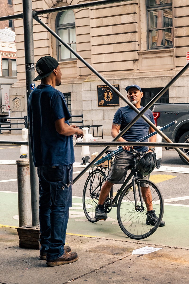 Two workers talking under sidewalk scaffolding, one holding a bicycle and boxes of fluorescent tubes