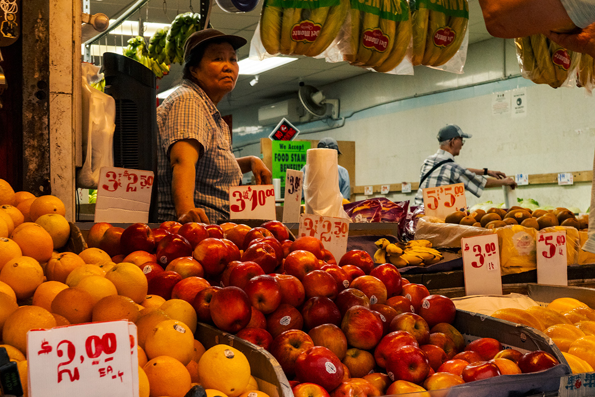 Woman working behind a fruit stand filled with oranges, apples, bananas, and handwritten price signs