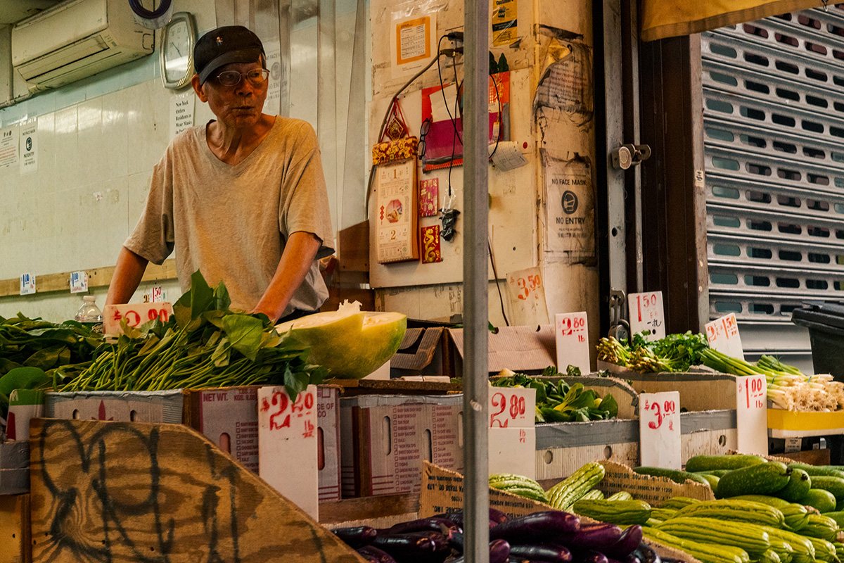 Man working behind a market display of leafy vegetables, melons, and handwritten price signs