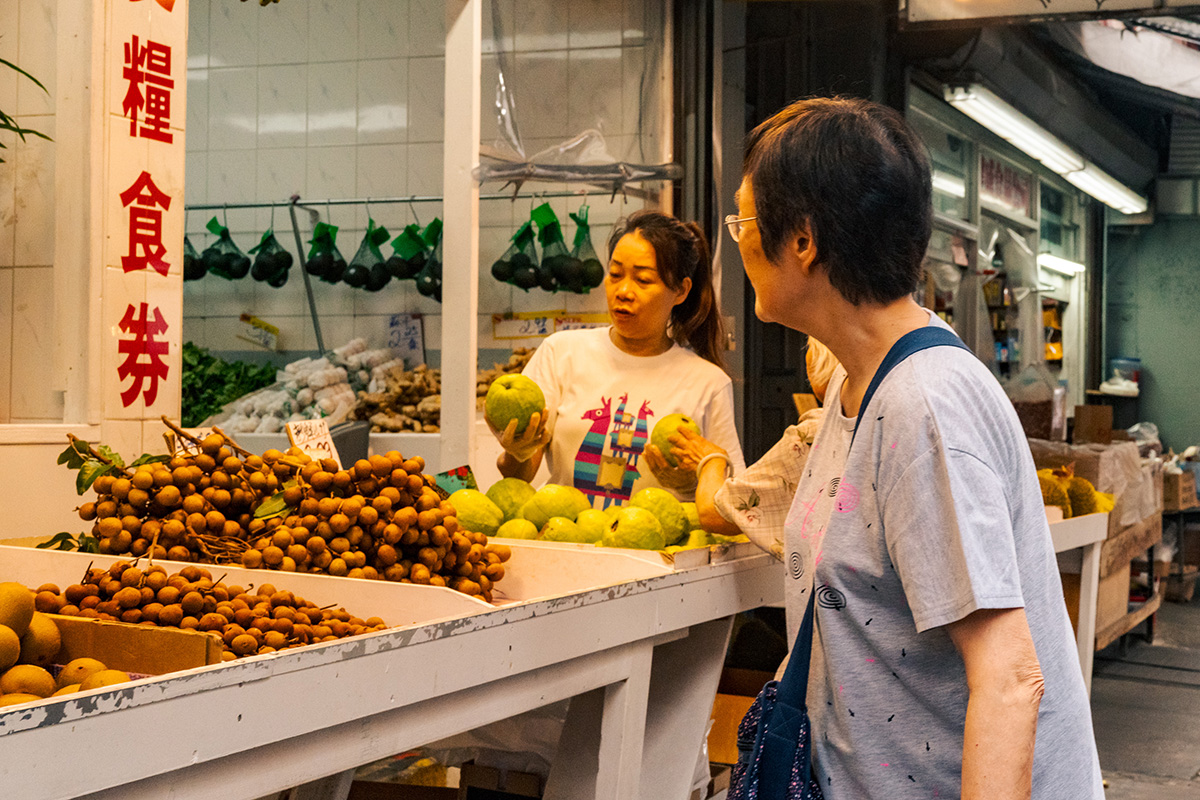 Fruit vendor holding two green fruits while speaking with a customer across the market counter