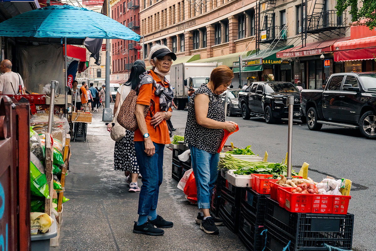 Two women beside an outdoor vegetable stand on a busy Chinatown street
