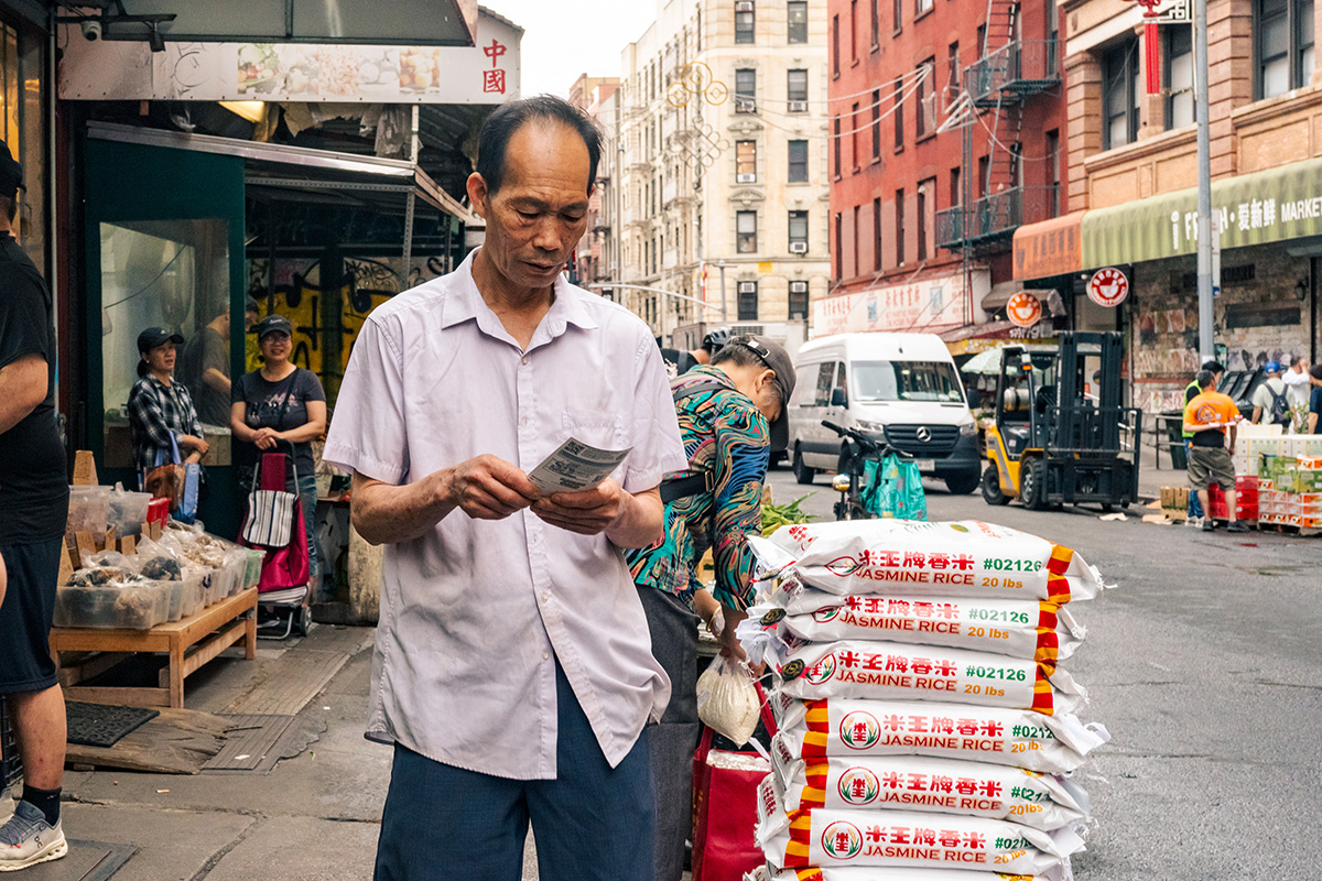 Man reading a lottery ticket beside a stack of jasmine rice bags on a Chinatown sidewalk