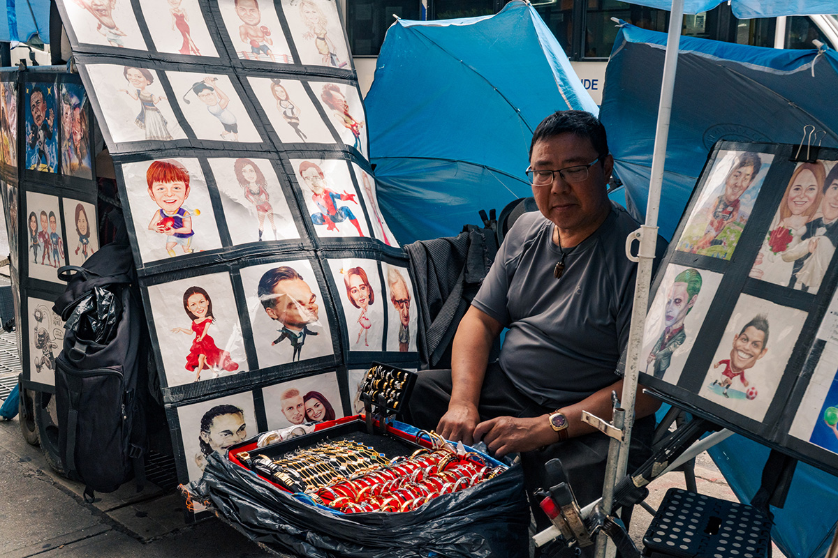 Seated street vendor selling bracelets and watches beneath displays of caricature portraits