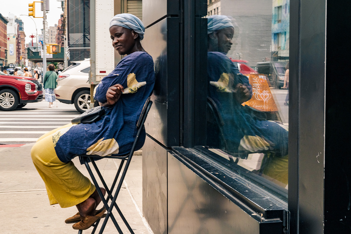 Seated woman in a blue and yellow dress reflected in a storefront window at a city intersection