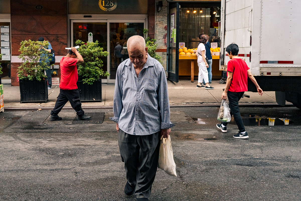 Older man carrying a shopping bag across a Chinatown street with pedestrians and a delivery truck behind him