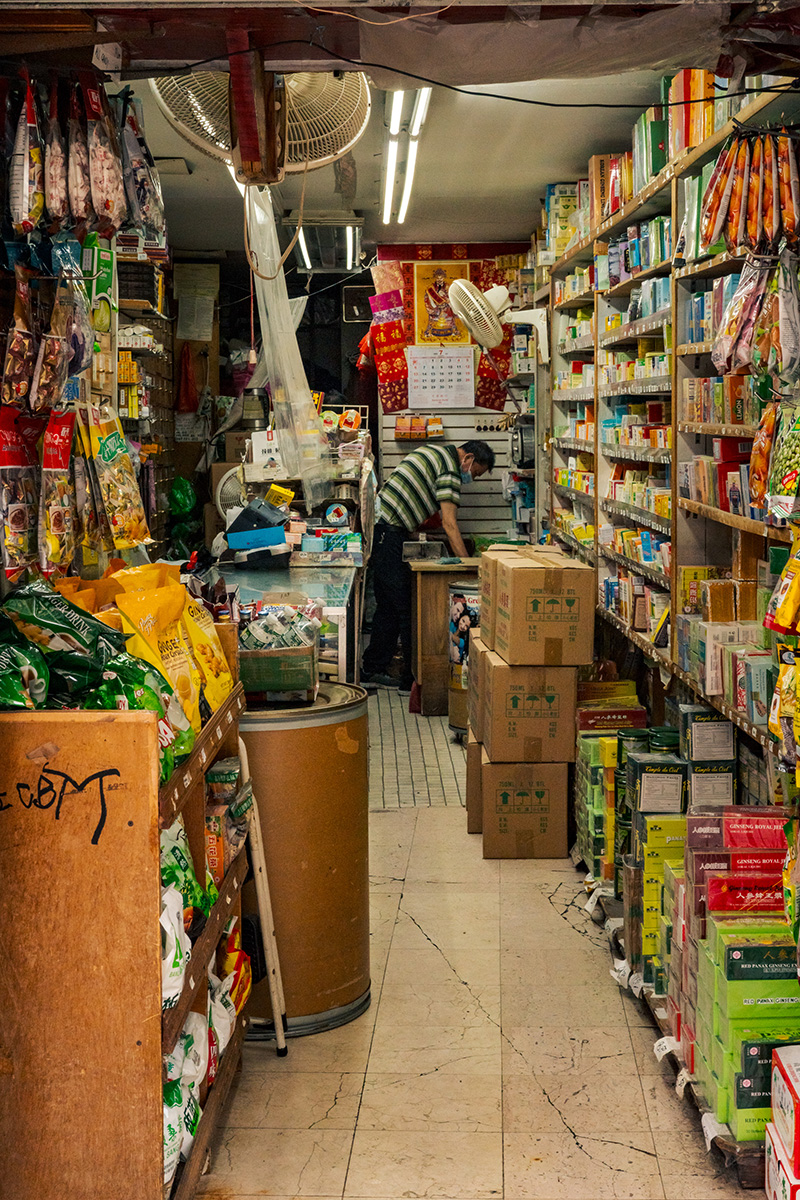 Masked shop worker bending over in a narrow market aisle lined with packaged goods and stacked boxes
