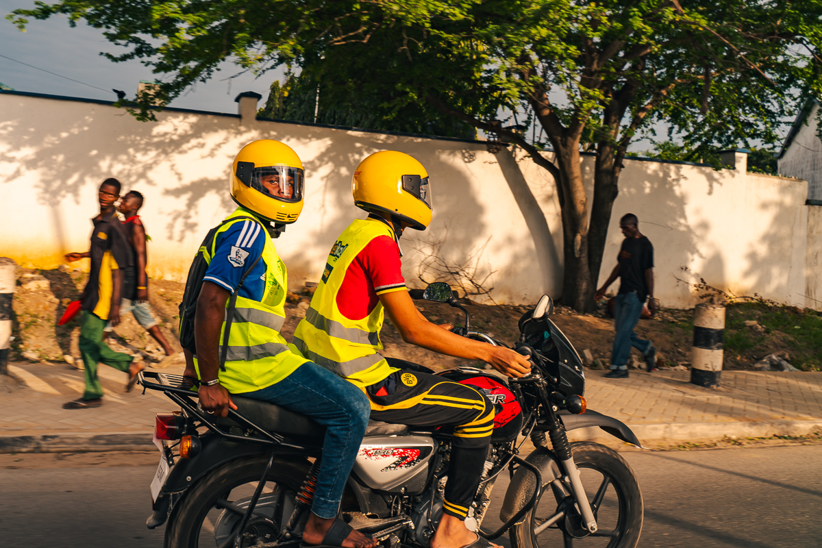 Two people in yellow helmets and reflective vests riding a motorcycle past pedestrians