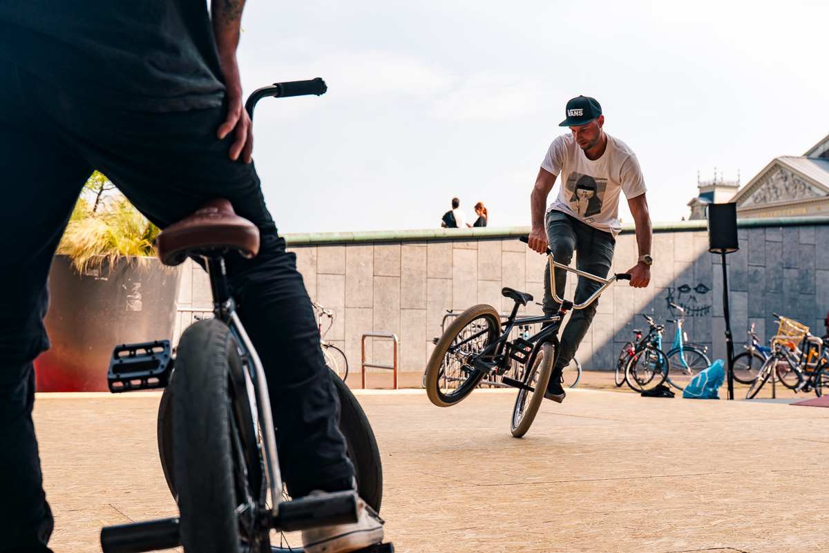 BMX rider balancing on one wheel, framed by another rider and bicycle in the foreground