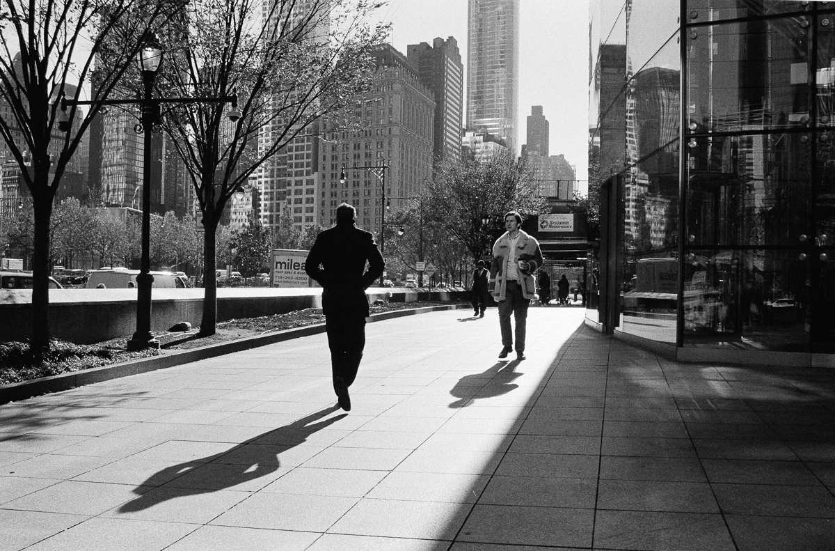 Two walkers casting long shadows across a sunlit city sidewalk, in black and white