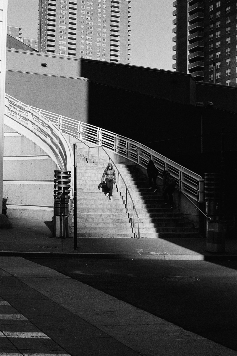 Person walking down sunlit stairs beside a curved ramp, with two people walking up the stairs in deep shadow.