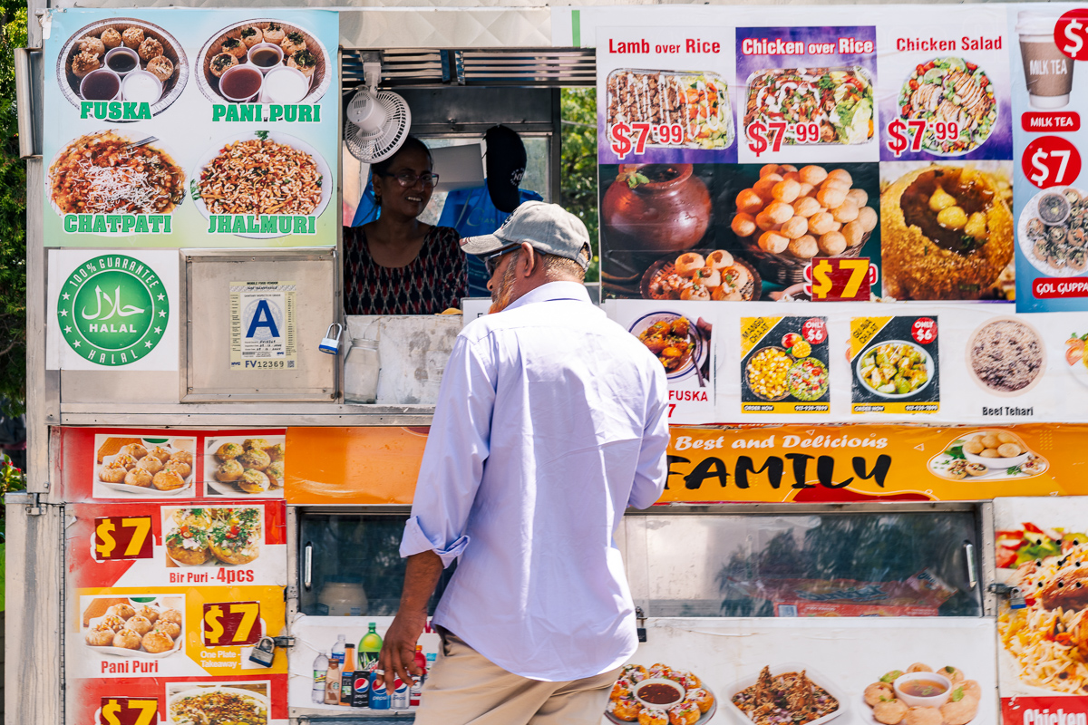 Customer facing a daytime food stand covered with colorful menus, with a vendor at the window