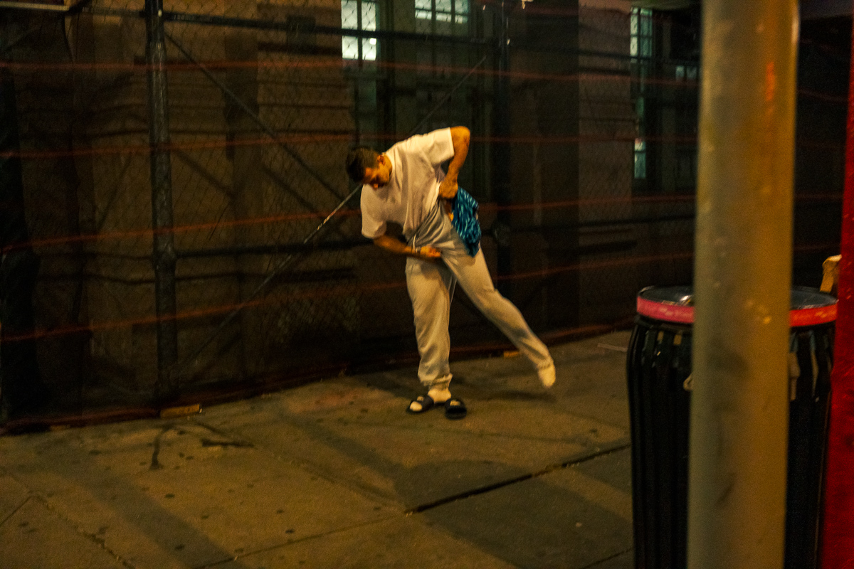 Man leaning forward while adjusting his pants on a dim sidewalk beneath scaffolding.