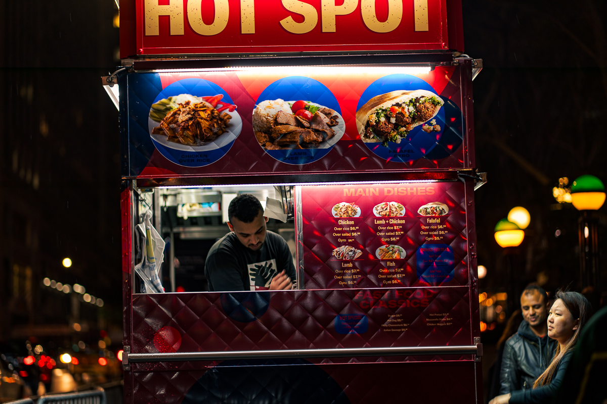 Illuminated Hot Spot food cart at night, with a vendor visible through the service window and two customers beside the cart.