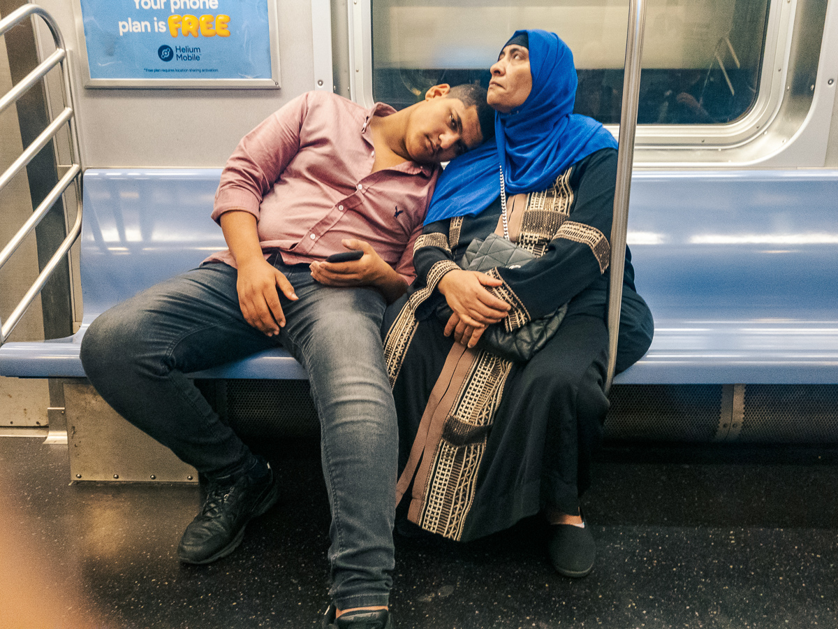 Two people seated together inside a subway car, one resting their head on the other's shoulder.